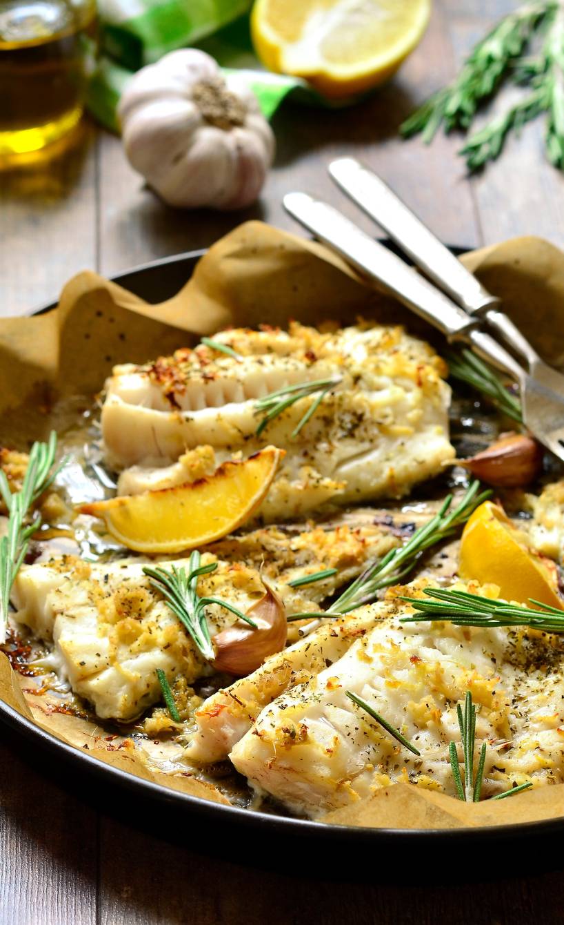 Kabeljau aus dem Ofen in einer Auflaufform garniert mit Zitronenspalten und Rosmarinzweigen auf einem Holztisch