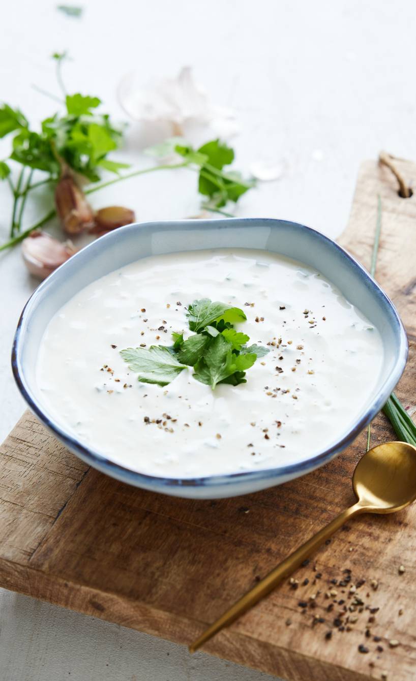 Knoblauch-Dip in einer wellenförmigen Schale auf einem Holzbrett garniert mit frischen Kräutern, daneben Knoblauchzehen