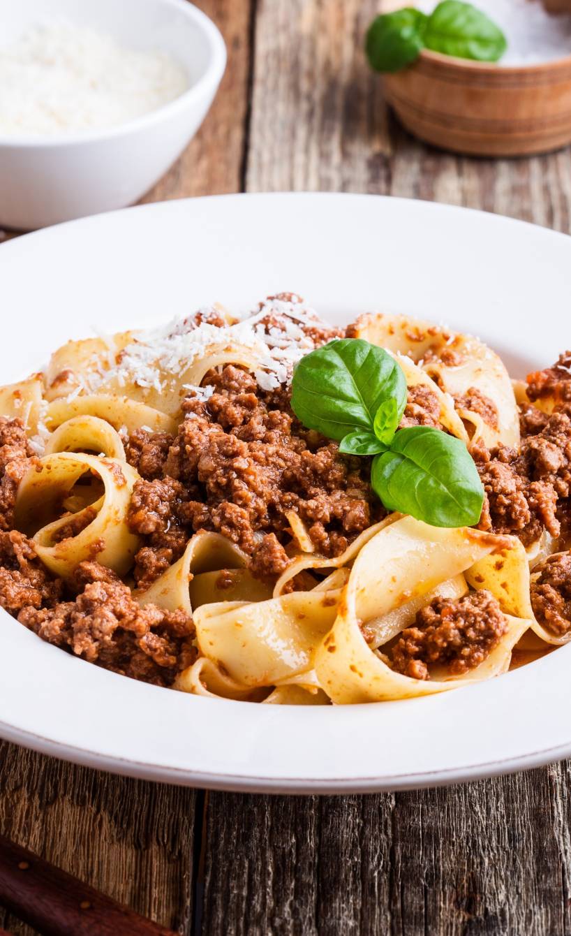 Pappardelle mit Bolognese aus Kalbshackfleisch auf einem tiefen weißen Teller, daneben Parmesan und Gewürze auf einem Holztisch