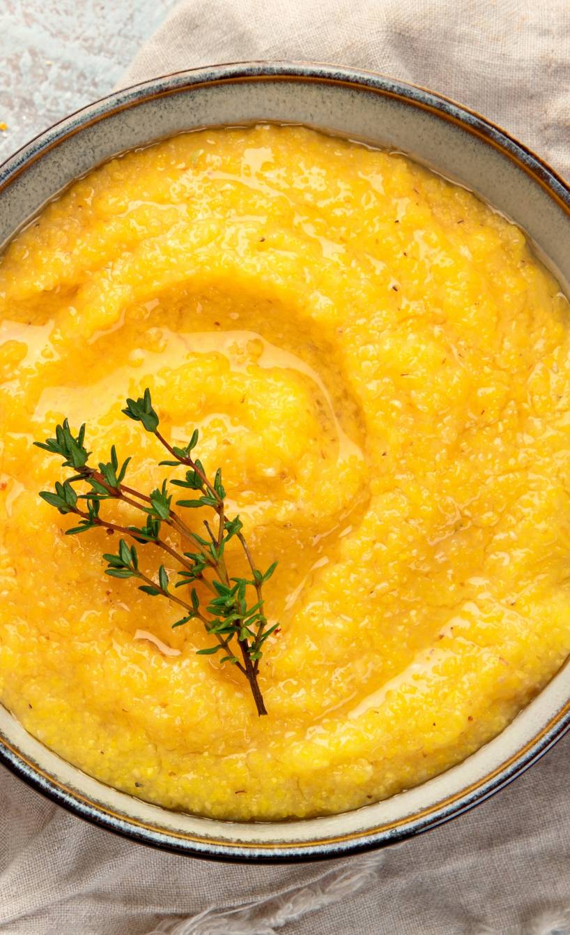 Goldgelbe Polenta von oben in einer Schüssel, darauf ein Zweig Gewürze, daneben Maisgrieß