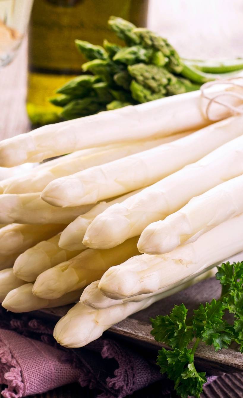 Päckchen von weißem und grünem Spargel auf einem Holztisch, dahinter eine Flasche Olivenöl und frische Kräuter