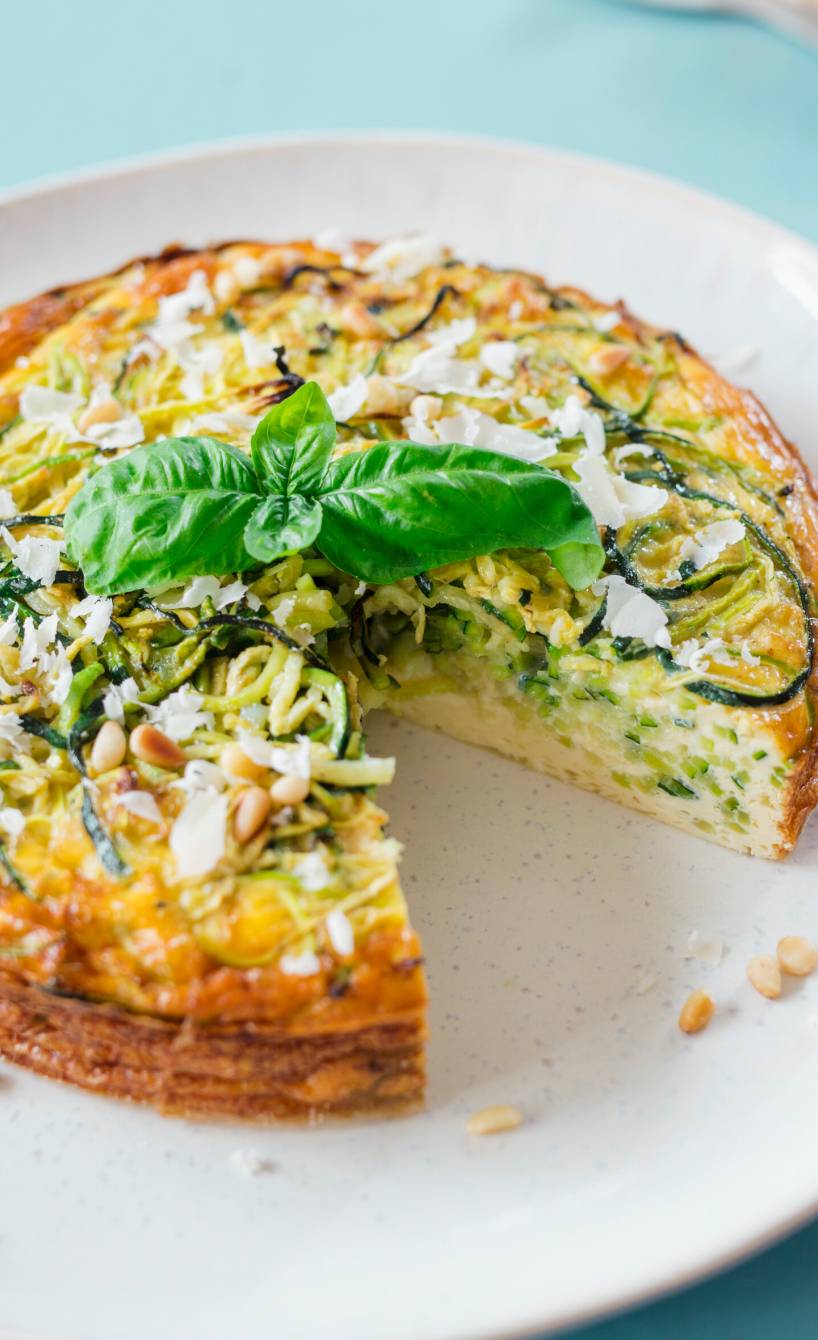 Runder Zucchini-Nudel-Kuchen angeschnitten auf einem Teller garniert mit Basilikum, daneben ein Stück Parmesan
