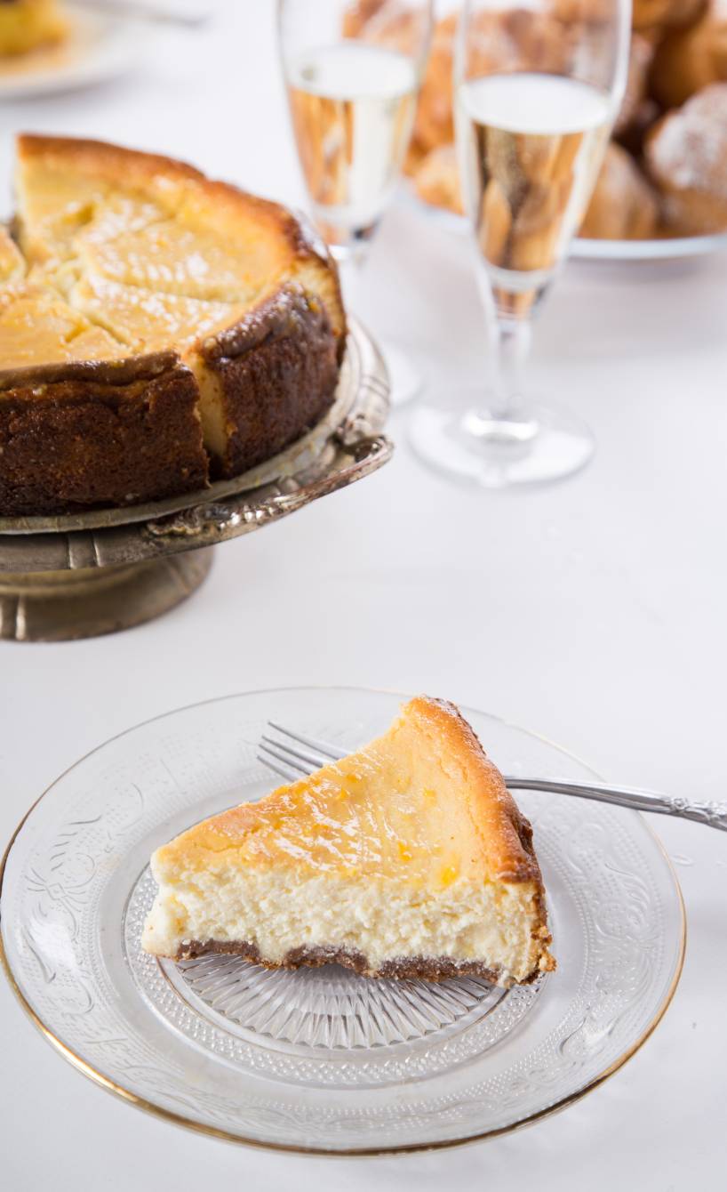 Ein Stück Ricotta-Cheesecake auf einem Kuchenteller, dahinter der restliche Kuchen auf einer Etagere