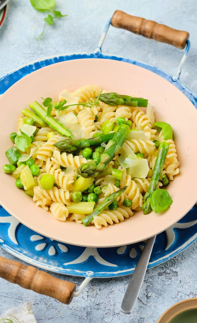 Pasta alla Primavera aus Fusilli, grünem Spargel und Erbsen in Sahnesauce auf einem Teller, darunter ein Platzteller mit Mosaik