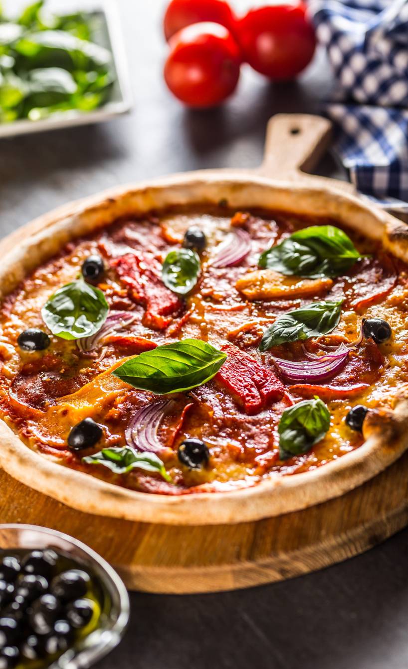 Runde Pizza Diavolo auf einem Holzbrett, daneben Zutaten wie Peperoni und Tomaten