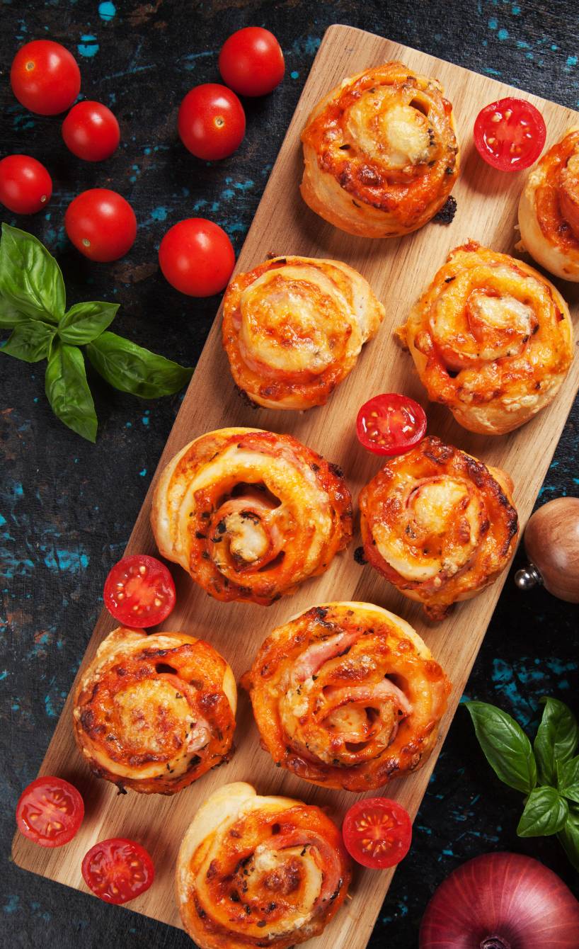 Neun Pizzaschnecken auf einem rechteckigen Holzbrett, daneben Tomaten auf einem Tisch