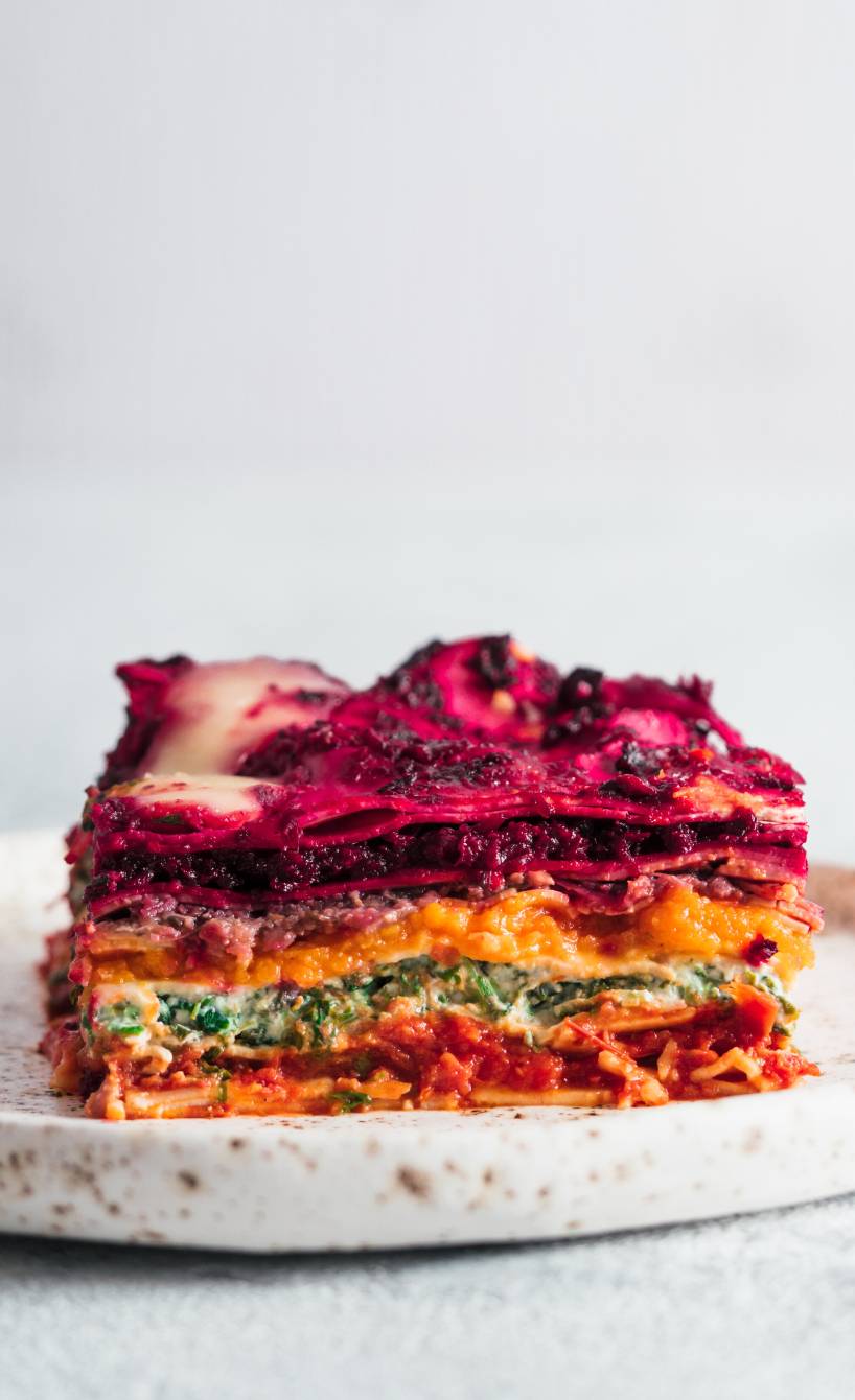 Ein Stück Regenbogen-Lasagne auf einem weißen Teller, bunte Gemüseschichten und Mozzarellakruste gut sichtbar