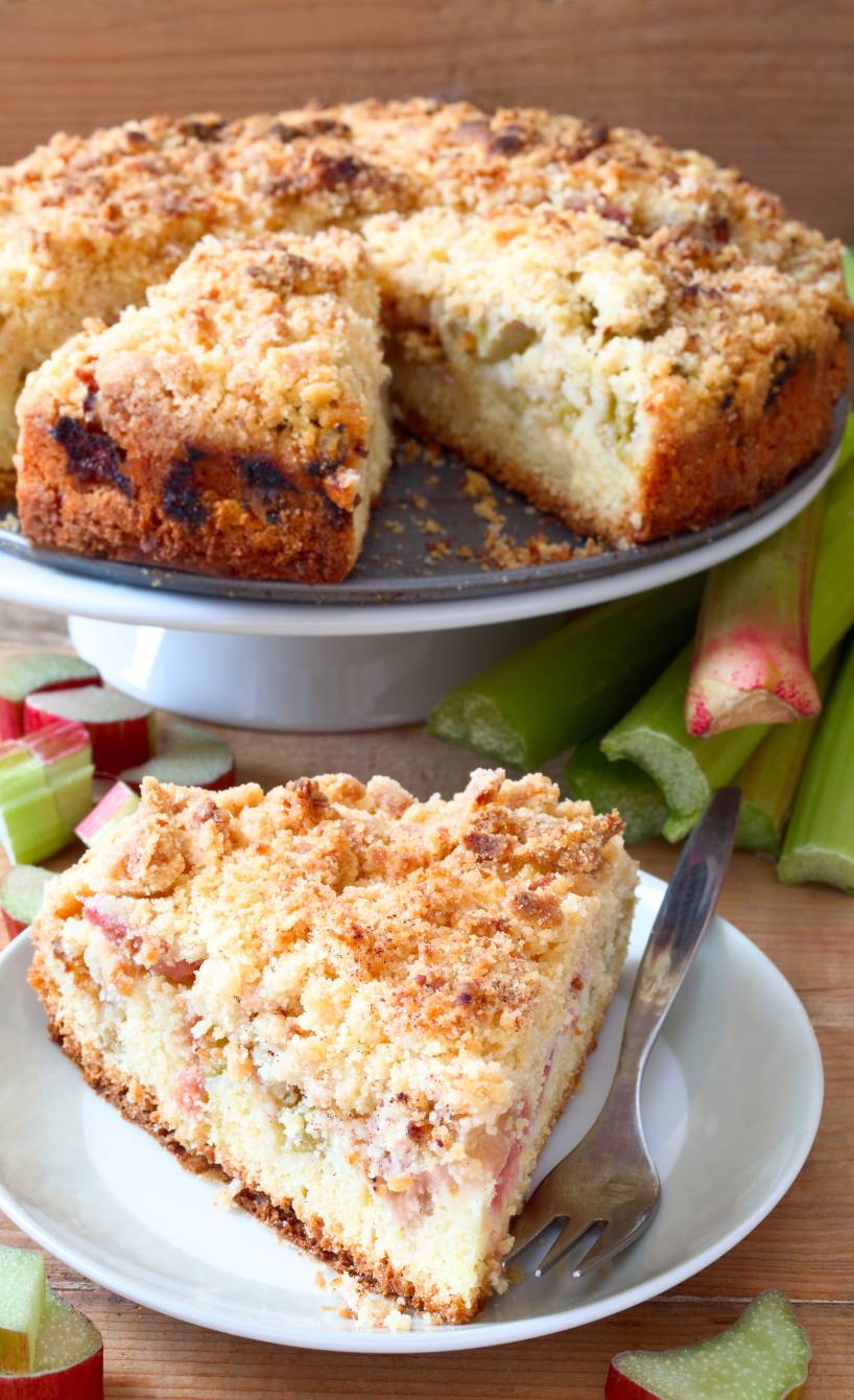 Runder Rhabarber-Streuselkuchen auf einer Kuchenplatte, davor ein Stück auf einem Teller