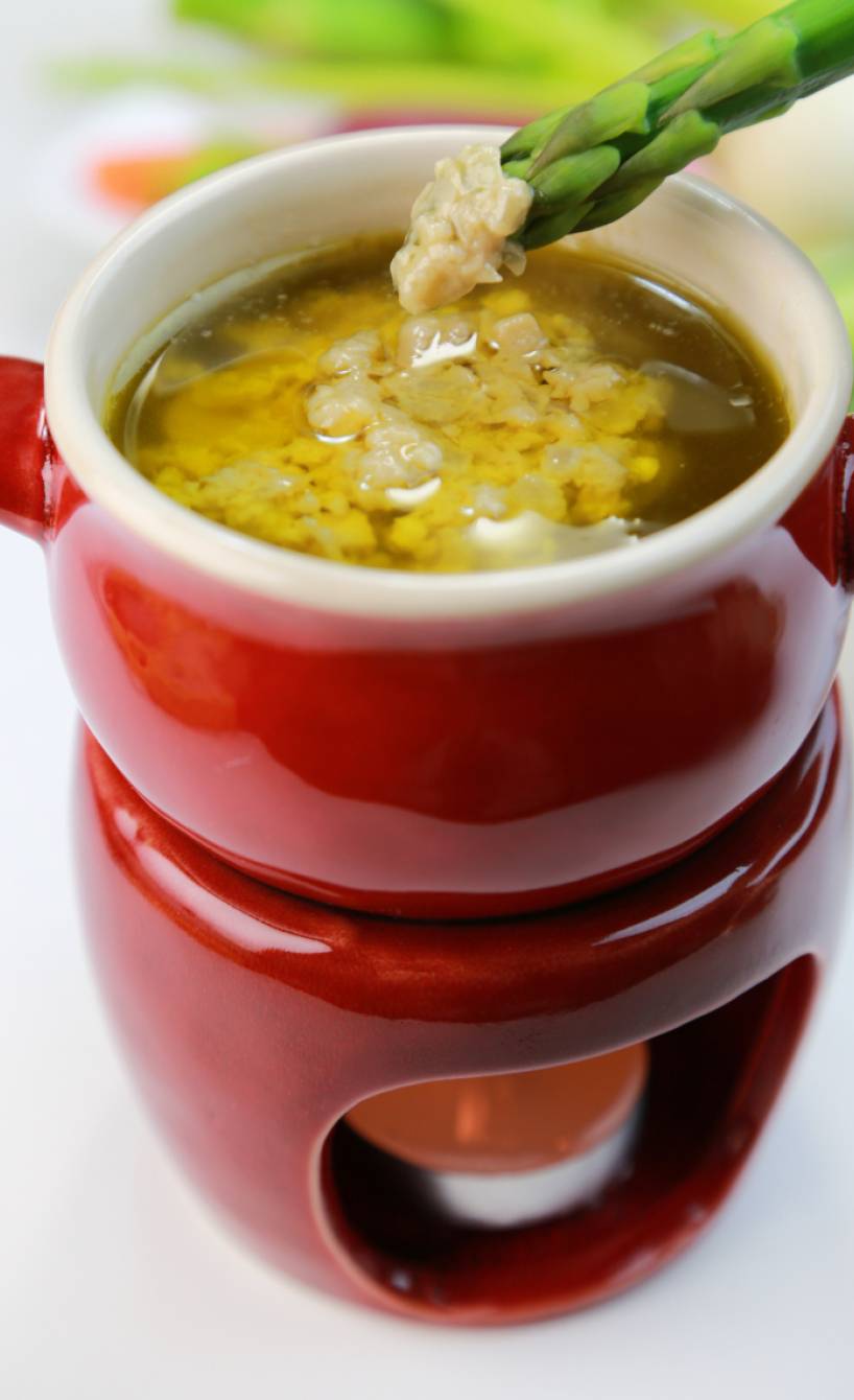 Bagna Cauda Sardellen-Knoblacuh-Sauce in einem Steinguttopf auf einem Stävchen, es wird grüner Spargel hineingedippt