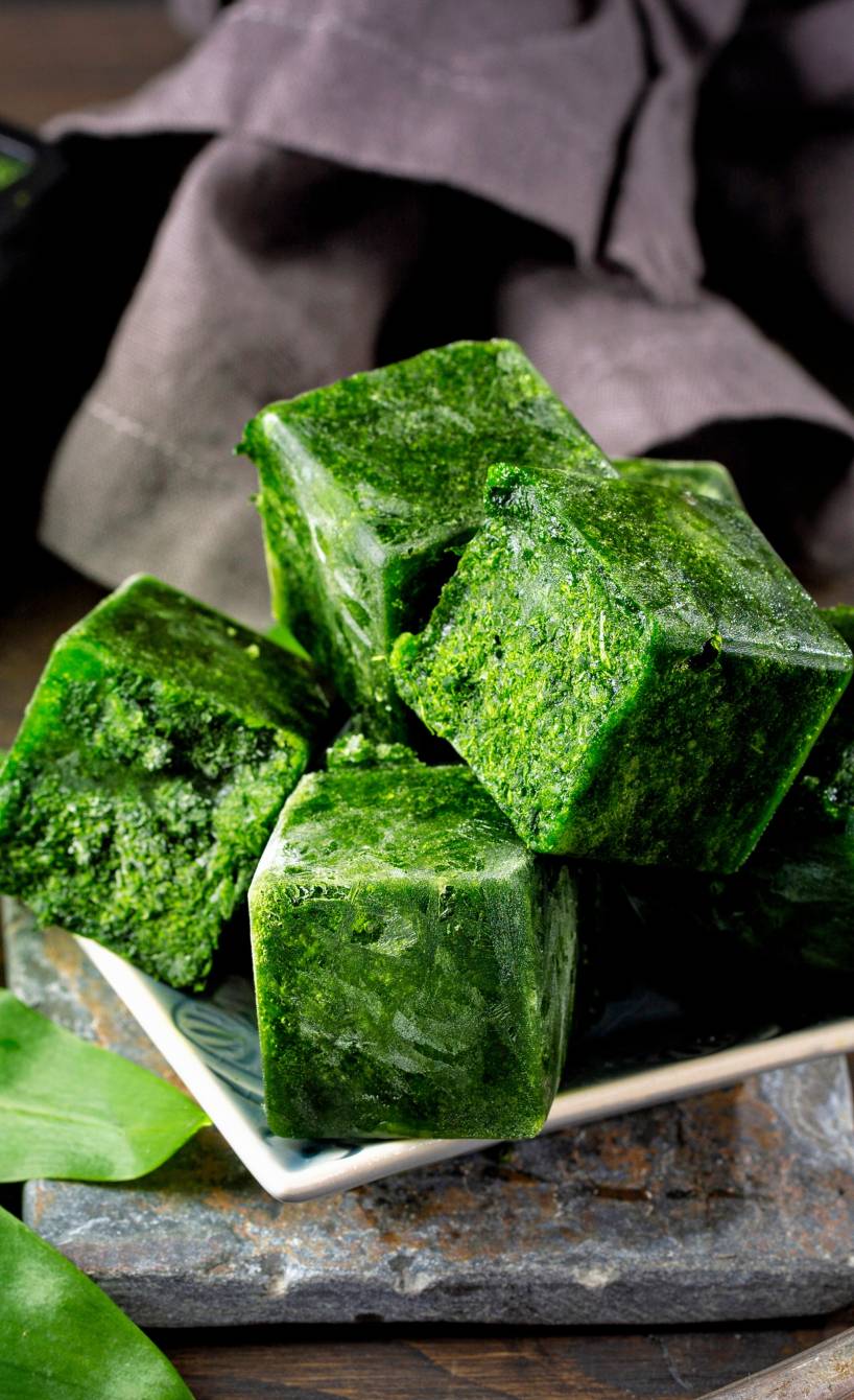 Würfel aus eingefrorenem Bärlauch auf einem Teller, dahinter eine Eiswürfelform und frischer Bärlauch