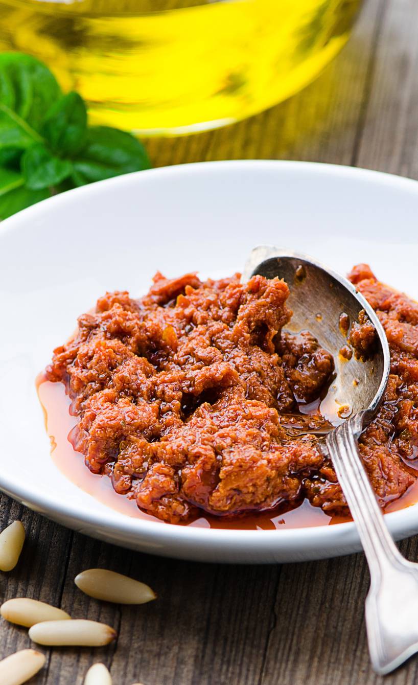 Selbst gemachtes Pesto rosso in einer Schale, daneben Pinienkerne und Olivenöl auf einem Holztisch