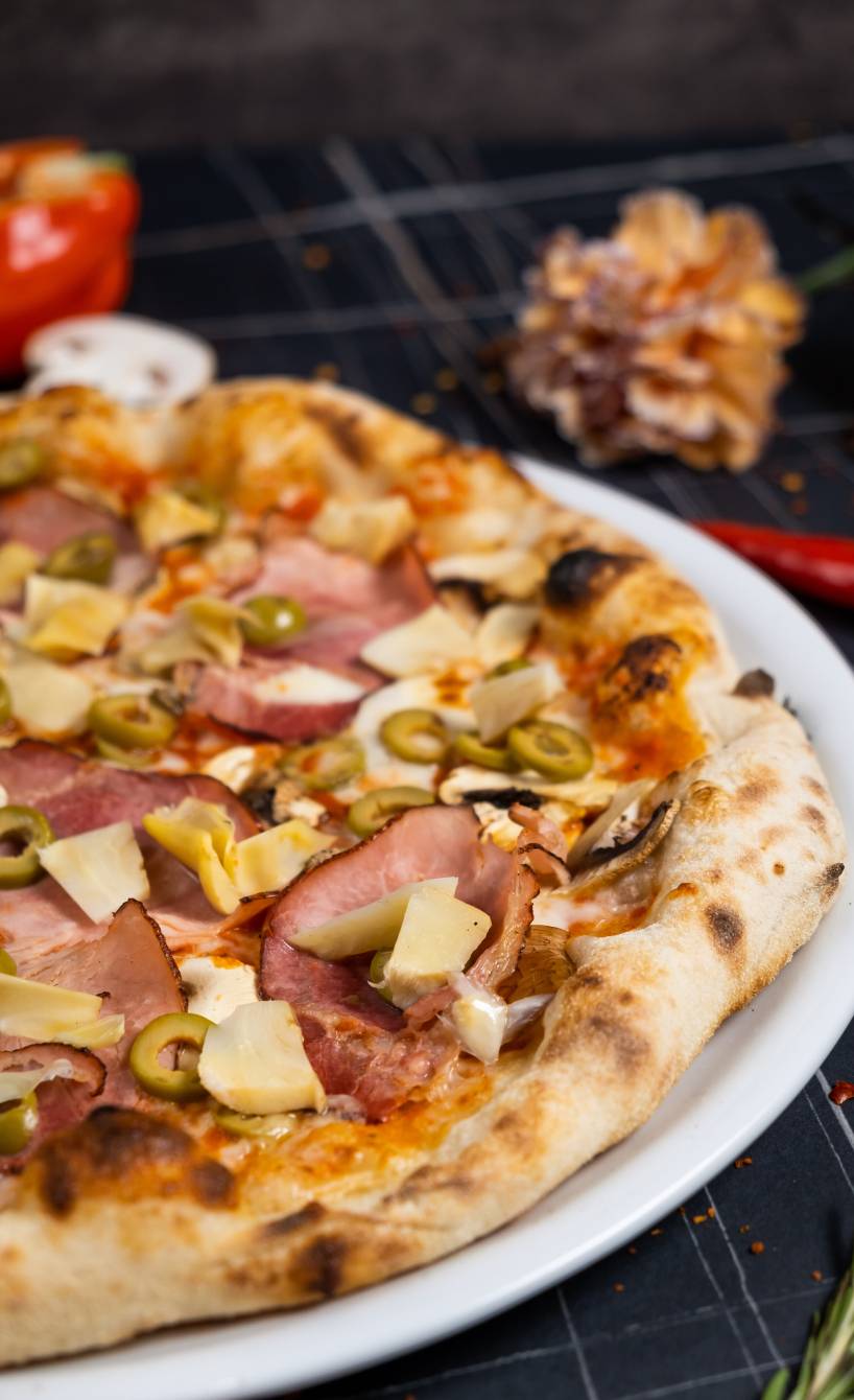 Pizza Capricciosa auf einem Pizzateller belegt mit Mozzarella, Artischocken, Champignons, Schinken und Oliven vor einem Backofen auf einem Tisch