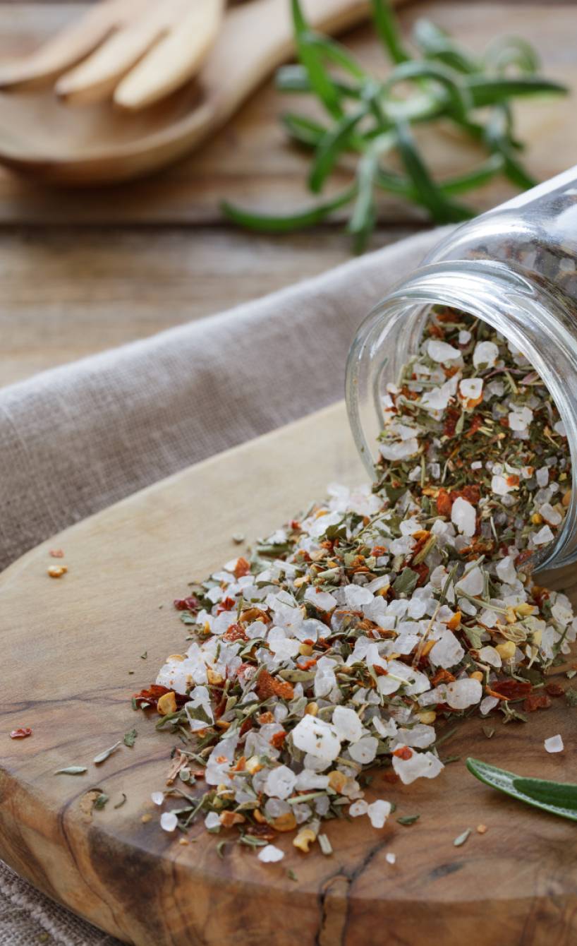Selbst gemachte Bruschetta-Gewürzmischung in einem Gewürzglas, das umgekippt auf einem Holzbrett liegt, darauf etwas von dem Gewürz