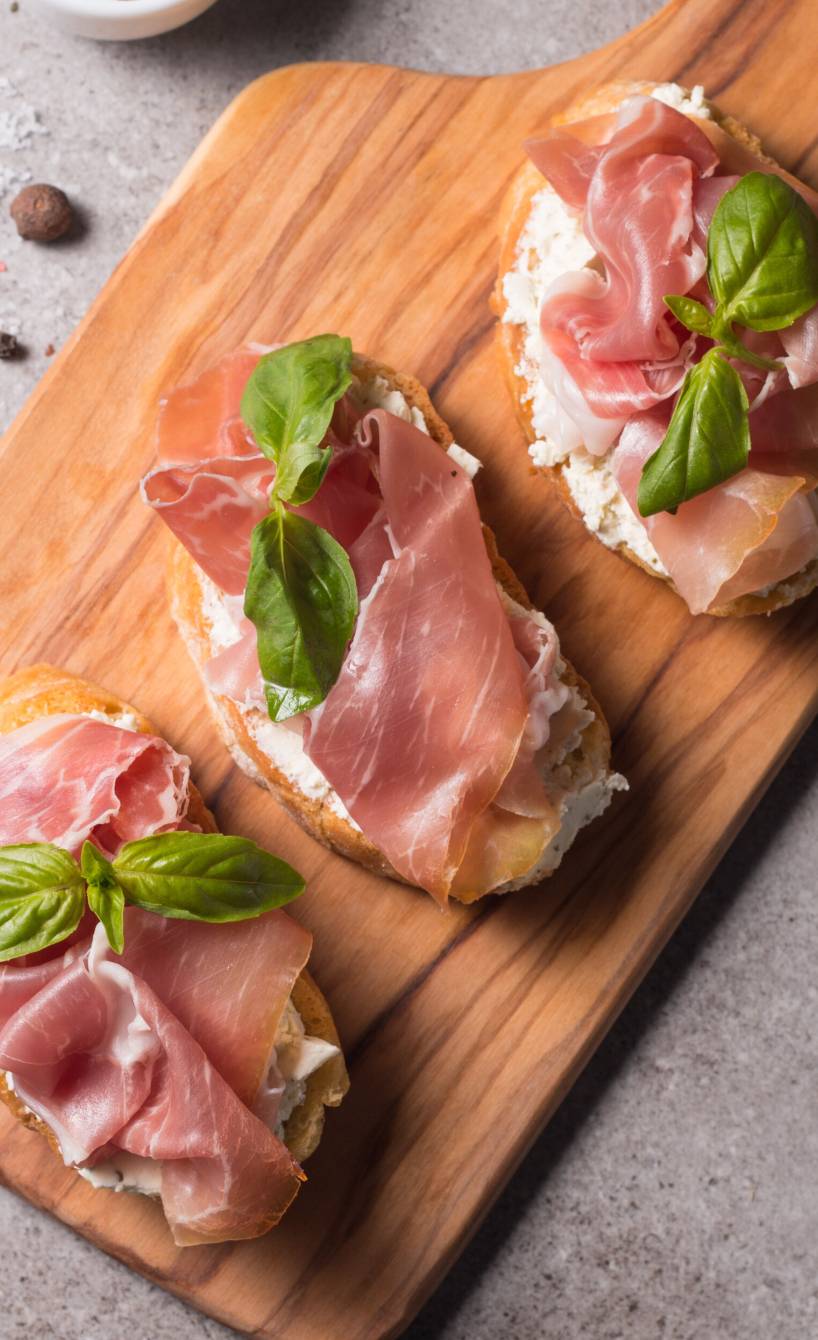 Drei Bruschettas mit Frischkäse und Schinken belegt auf einem Holzbrett garniert mit frischem Basilikum