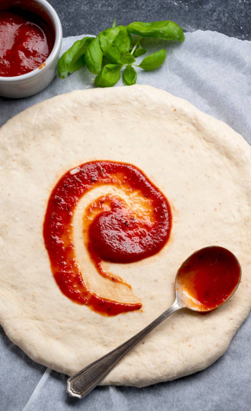 Glutenfreier Pizzateig ausgerollt als runder Pizzaboden wird mit Tomatensauce bestrichen, daneben frischer Basilikum