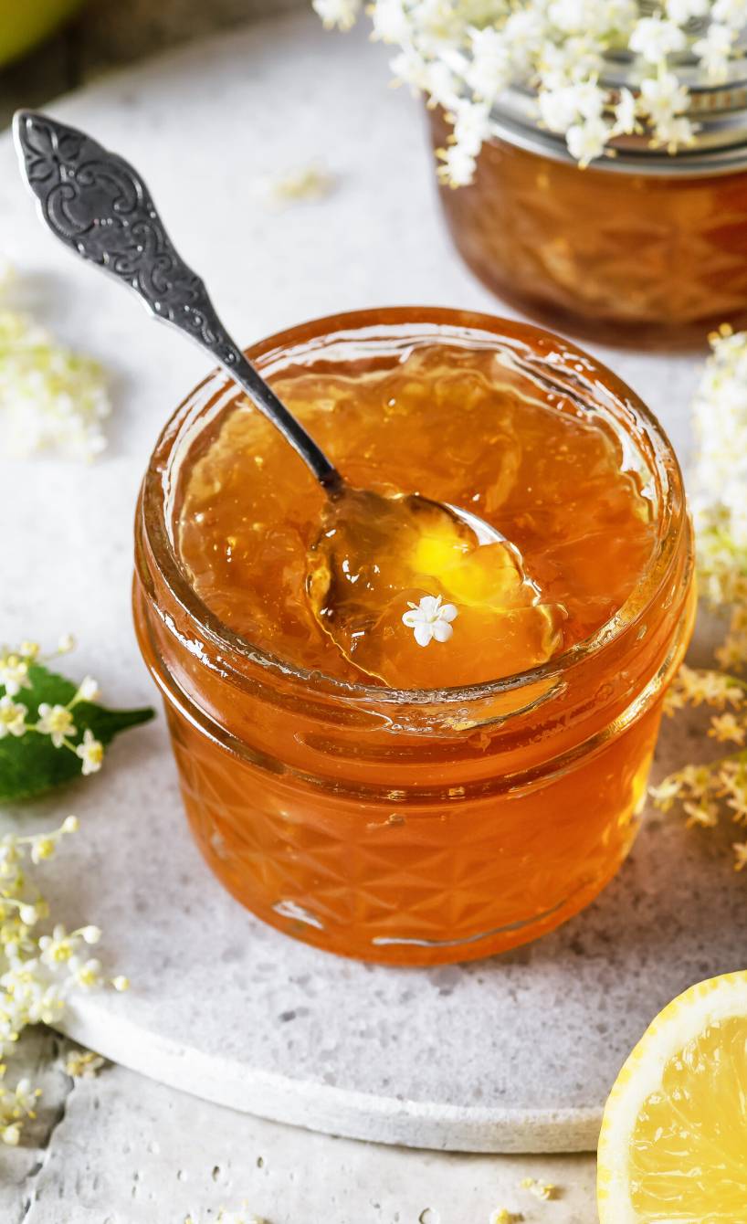 Selbst gemachtes Holunderblütengelee in zwei Einmachgläsern auf einem Tisch, daneben Holunderblütenzweige und Zitronen