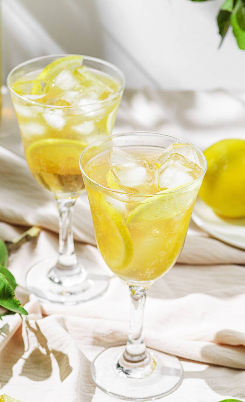 Limoncello Spritz mit Eiswürfeln in zwei Cocktailgläsern, daneben Zitronen, eine Flasche Limoncello und Zitronenmelisse auf einem Tisch