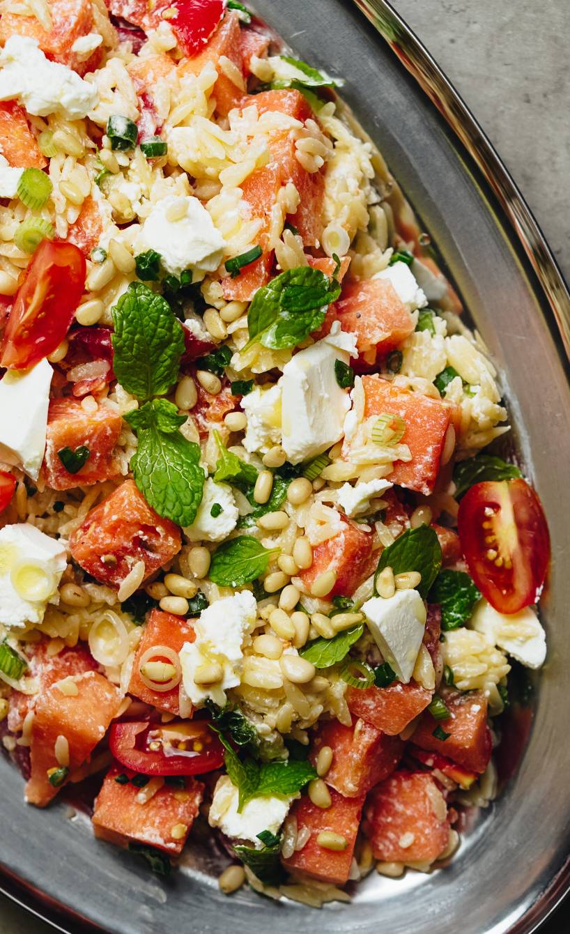 Orzo-Salat mit Feta und Wassermelone auf einem ovalen Teller getoppt von Pinienkernen und frischer Minze