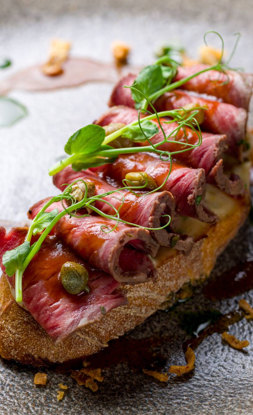 Bruschetta mit Roastbeef-Scheiben belegt, darauf Honig-Balasamico-Sauce, Kapern und frische Kräutern
