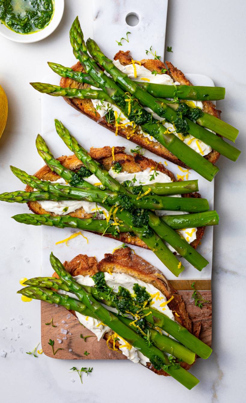 Drei Spargel-Ricotta-Bruschette auf einem Holzbrett getoppt von grünem PEsto und Zitronenabrieb