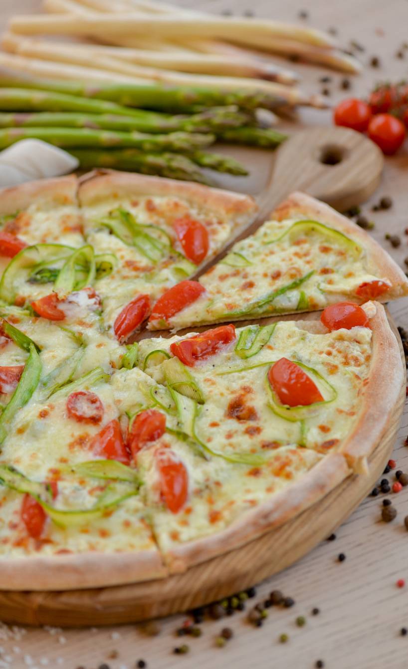 Spargelpizza mit weißem und grünem Spargel, Kirschtomaten Crème fraîche bestrichen angeschnitten auf einem Pizzabrett