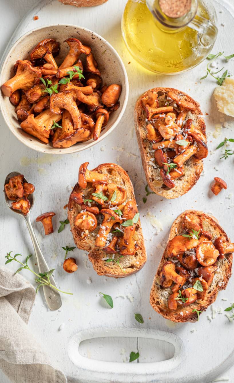 Drei Steinpilz-Bruschette auf einem Teller, daneben eine Schale mit Gebratenen Pilzen und Olivenöl auf einem Tisch