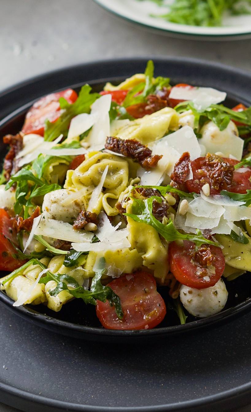 Tortellini-Salat mit Tomaten und Mozzarella auf einem Teller, daneben Rucola und Parmesan