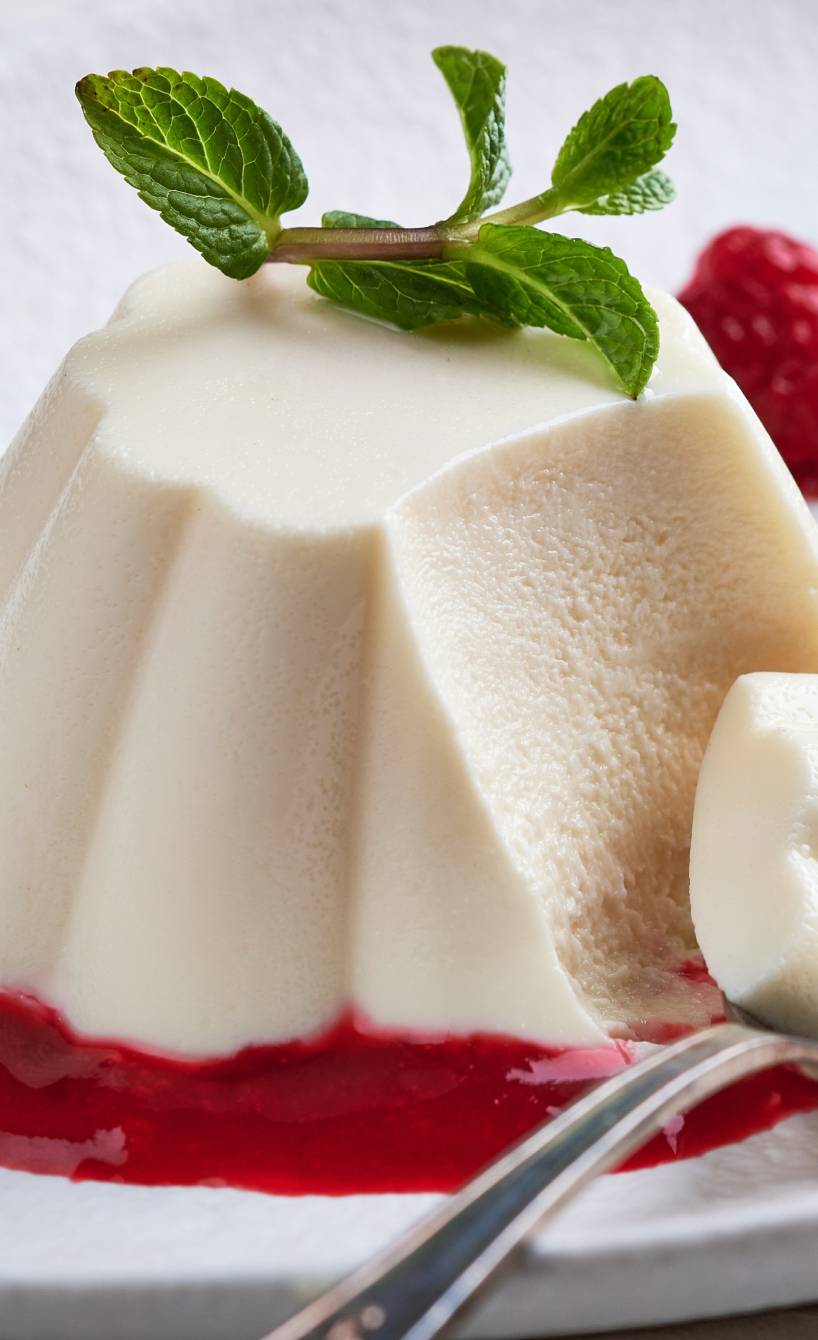 Panna Cotta auf einem Spiegel aus Himbeersoße, daneben frische Himbeeren und Minzblätter