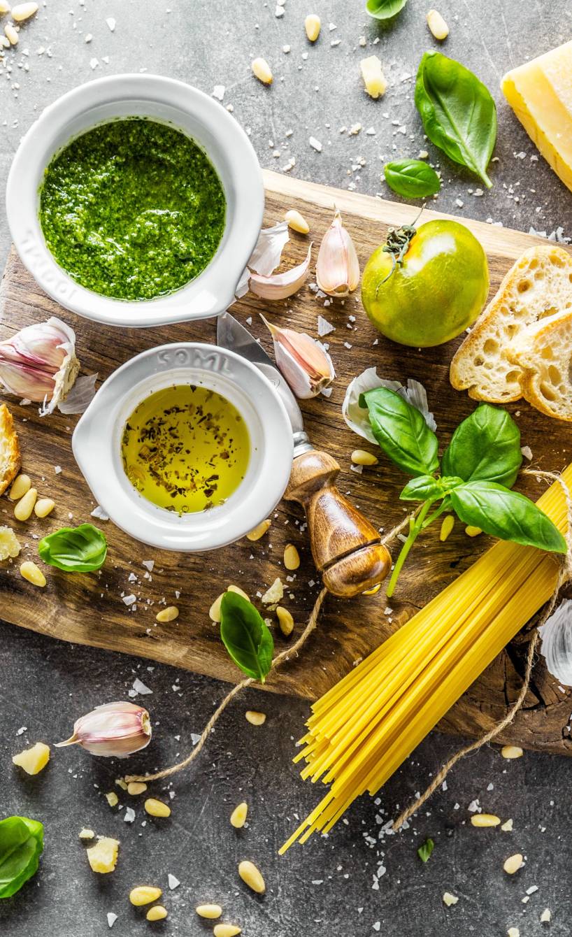 Grünes Pesto, Pestozutaten, ungekochte Spaghetti und Ciabatta von oben auf einem Holzbrett