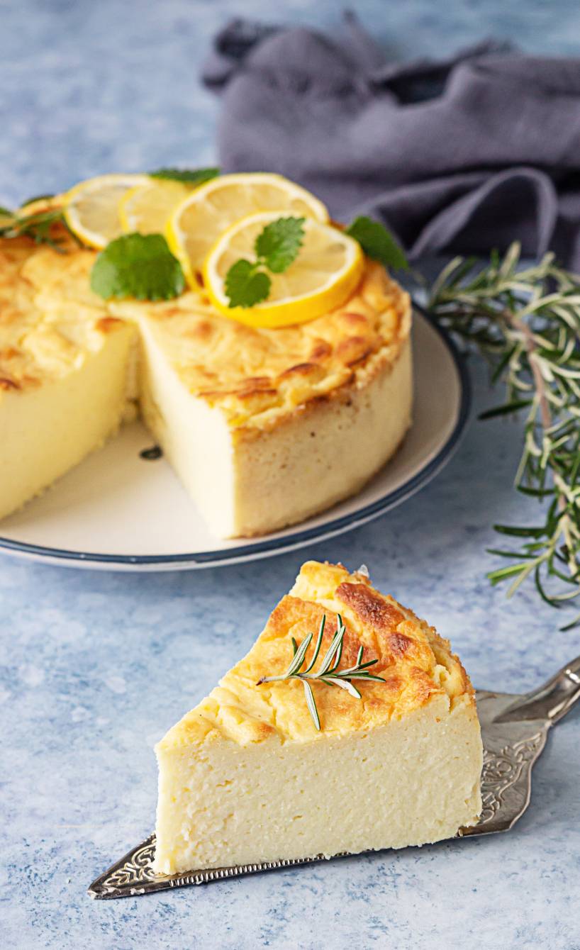 Zitronen-Ricotta-Kuchen (Torta di Ricotta e Limone) angeschnitten auf einer Kuchenplatte garniert mit Zireonenscheiben, Minzblättern und Rosmarin