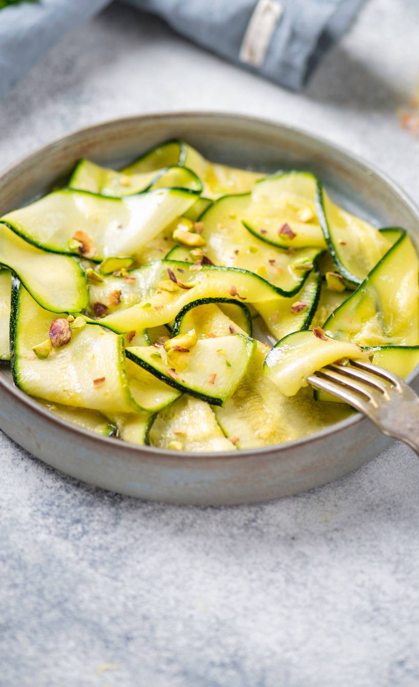 Rohes Zucchini Carpaccio in einer Schale getoppt von Parmesan und Pinienkernen, daneben eine Karaffe mit Olivenöl