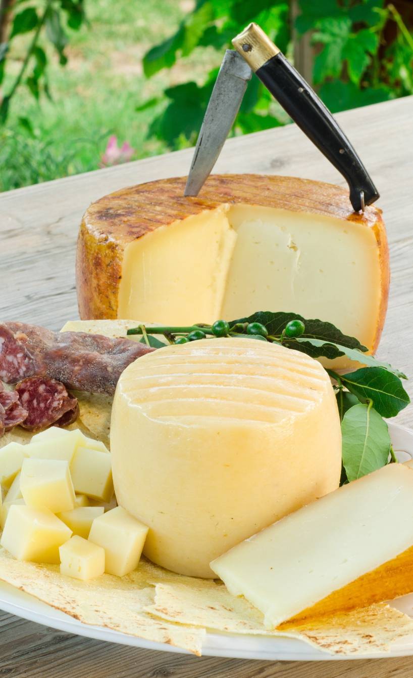 Platte mit einem runden, angeschnittenen Pecorino-Käselaib, daneben Salami und ein Glas Rotwein auf einem Holztisch