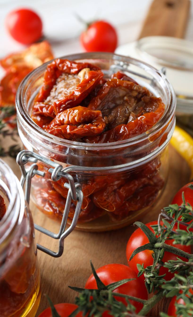 Eingelegte getrocknete Tomaten in Öl in einem Weckglas, daneben Knoblauch, frische Tomaten und Kräuter