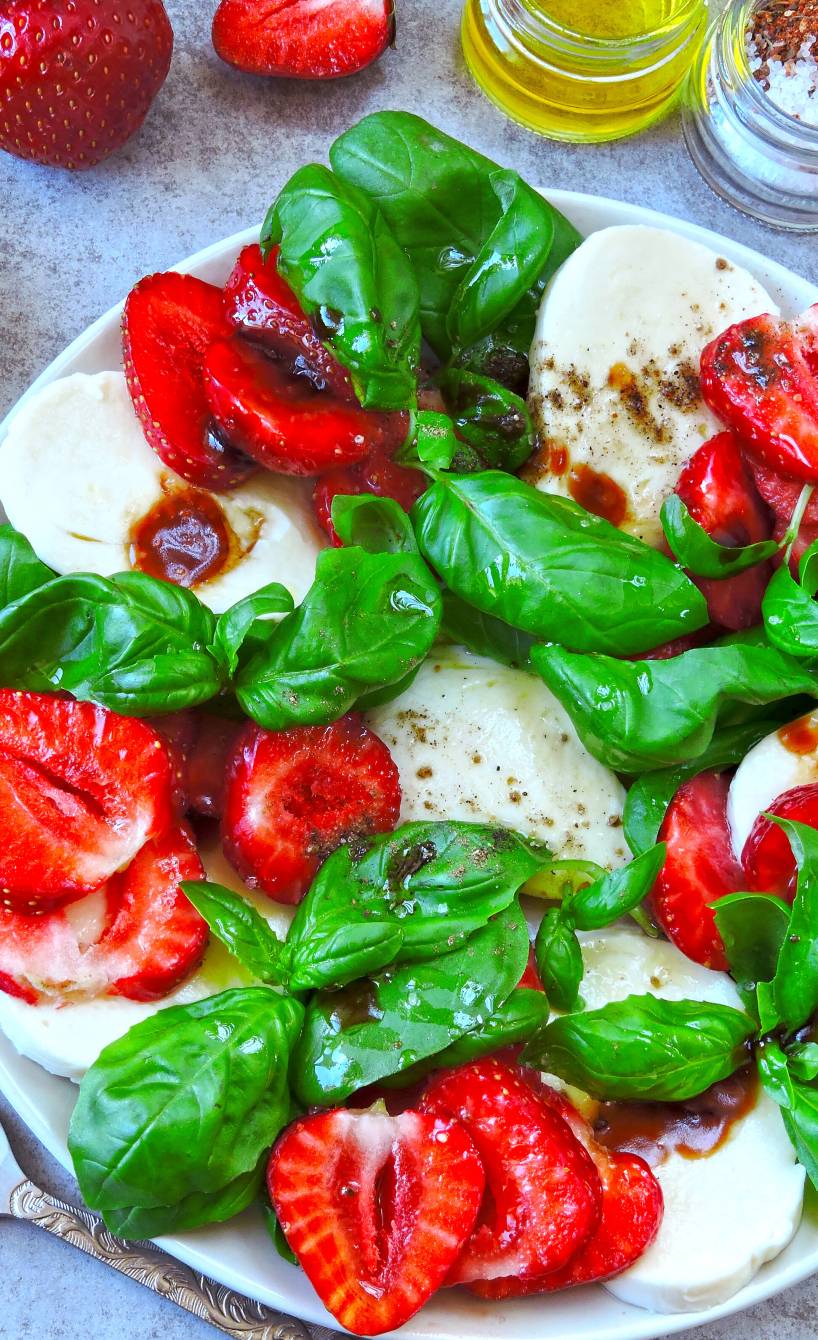 Erdbeer-Caprese angerichtet auf einem Teller mit Mozzarellascheiben und Basilikum, daneben Olivenöl