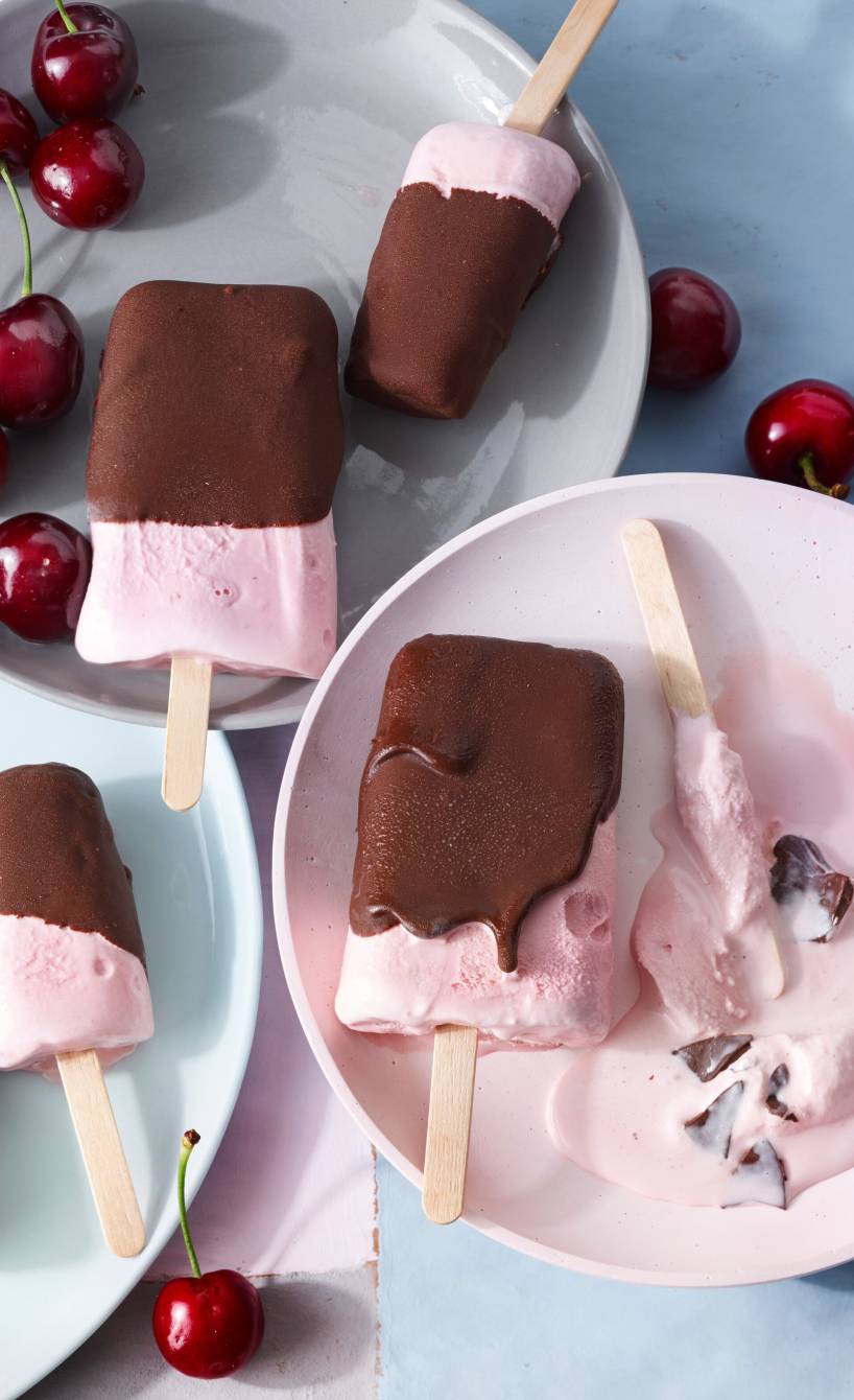 Selbstgemachtes Kirsch-Joghurt-Eis am Stiel mit Schokoglasur verteilt auf mehrere Teller, daneben frische Kirschen