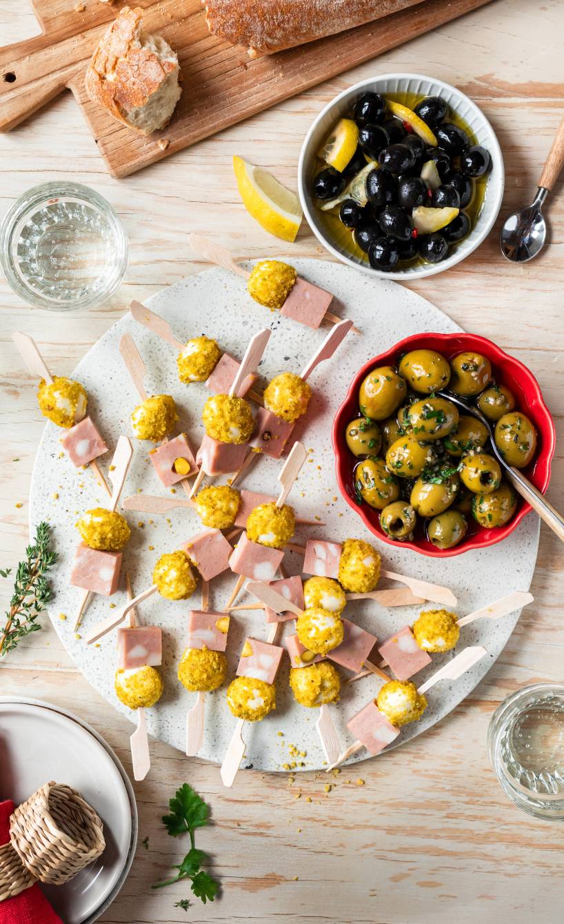 Mortadella-Mozzarella-Spieße auf einem runden Brett, daneben Oliven und Ciabatta