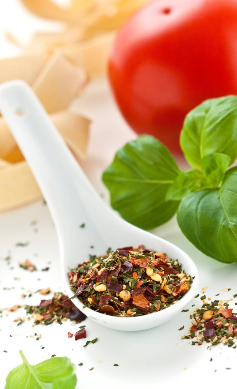 Selbst gemachtes Pizzagewürz auf einem Porzellanlöffel, daneben frischer Basilikum und Tomaten auf einem Tisch