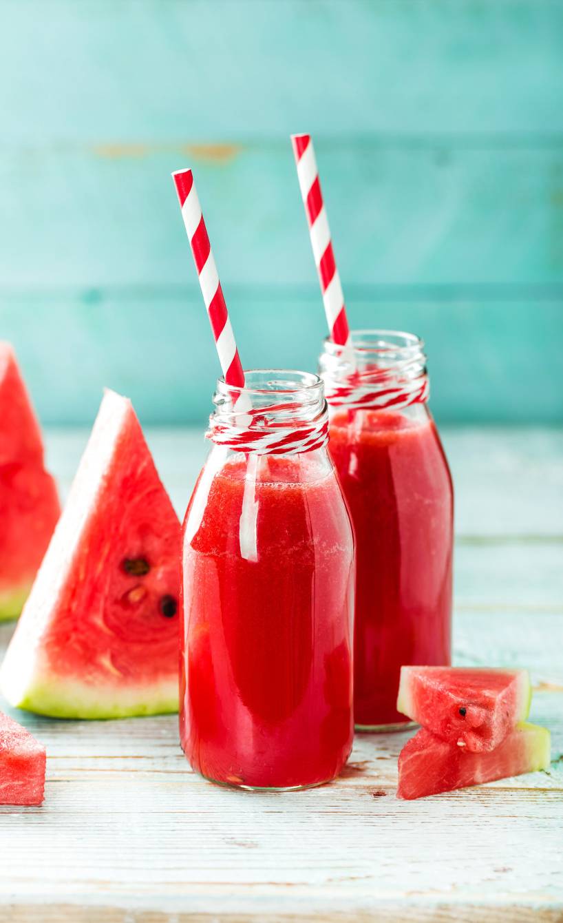 Wassermelonendrink in zwei Glasflaschen mit Strohhalmen, daneben Melonenstücke auf einem Tisch