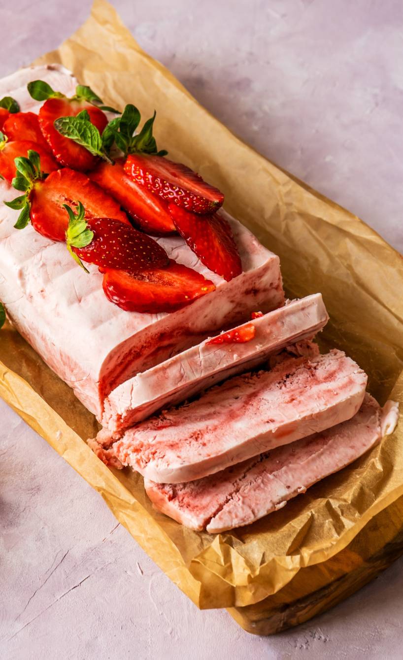 Selbst gemachtes Semifreddo angeschnitten auf einem Stück Backpapier getoppt von frischen Erdbeeren, daneben Erdbeersoße