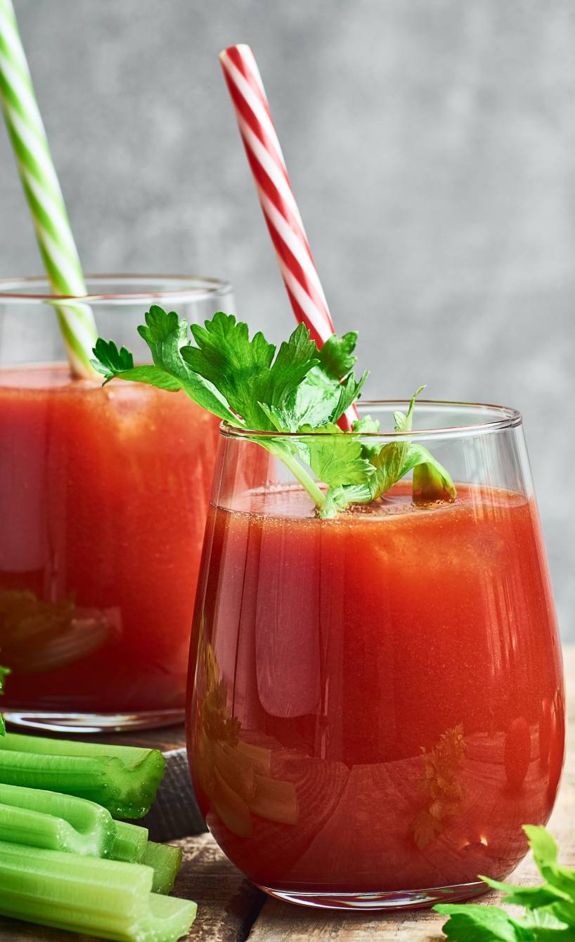 Getränk auf Tomatensaftbasis in zwei Gläsern auf einem Holztisch, daneben frische Tomaten, Sellerie und Kräuter