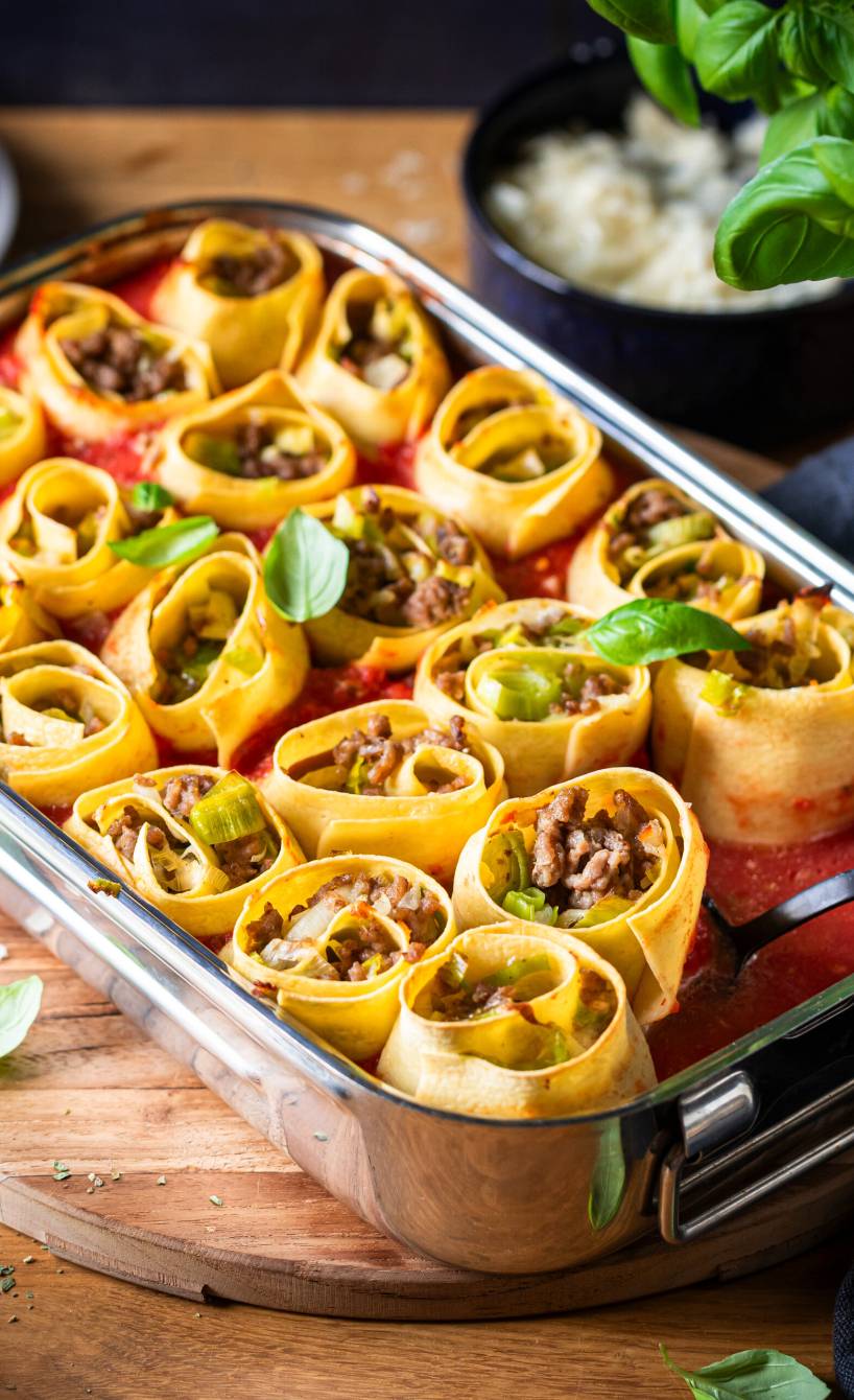Eine Auflaufform mit gebackenen Pastaschnecken gefüllt mit Lauch und Hackfleisch, daneben frischer Basilikum