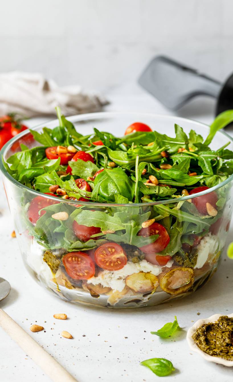 Italienischer Katoffelsalat mit Tomaten, Mozzarella und Rucola geschichtet in einer Glasschüssel, daneben grünes Pesto auf einem Tisch