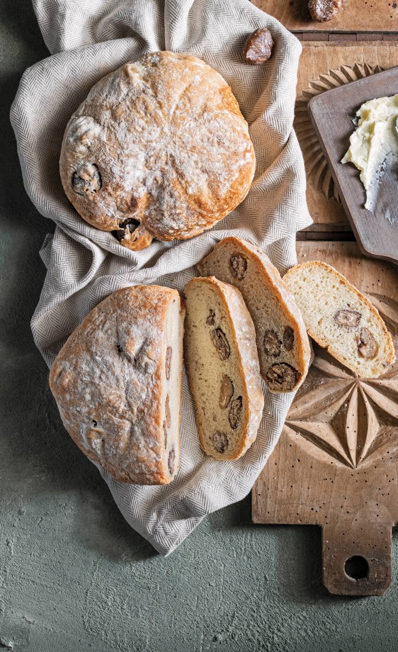 Zwei Laibe Maronenenbrot auf einem Tisch, eines angeschnitten, daneben gemusterte Holzbretter 