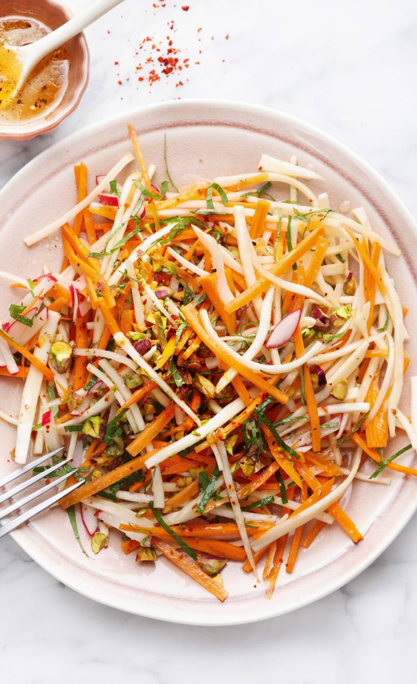 Roher Karottensalat mit Pastinaken, Radieschen und Pistazien auf einem Teller, daneben Orangendressing