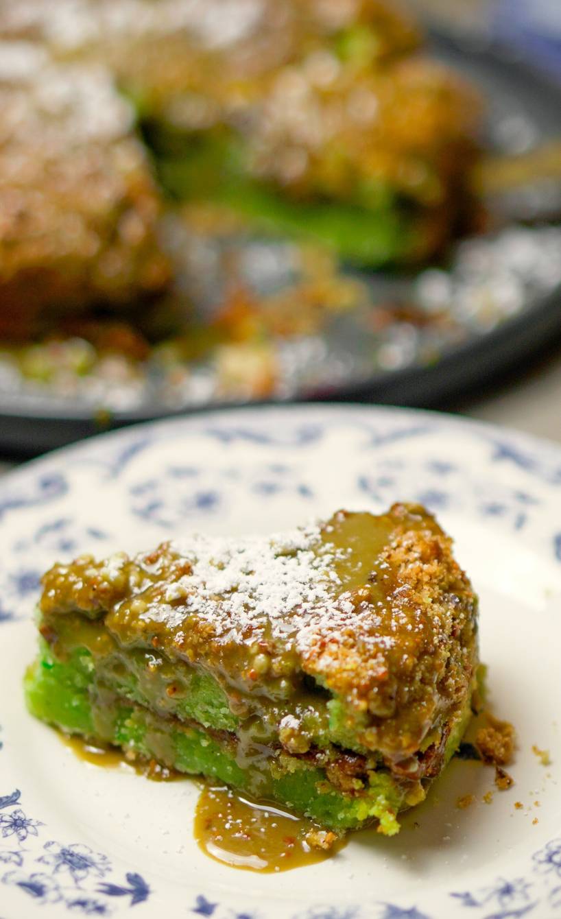 Ein Stück Pistazienkuchen auf einem Teller, dahinter der restliche Kuchen auf einer Kuchenplatte