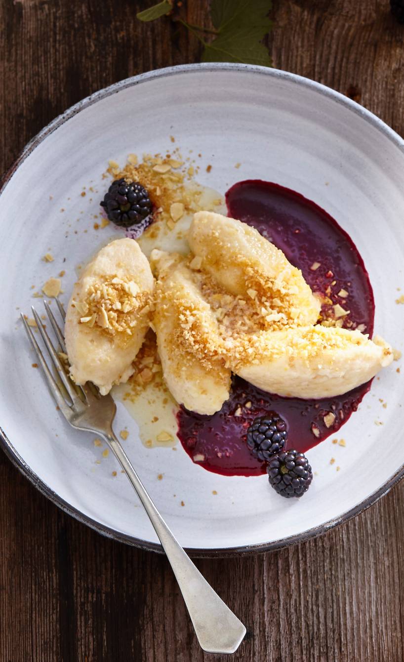 Vier Hüttenkäse-Grießnocken auf einem Dessertteller mit Brombeersauce bestreut mit Mandel-Bröseln