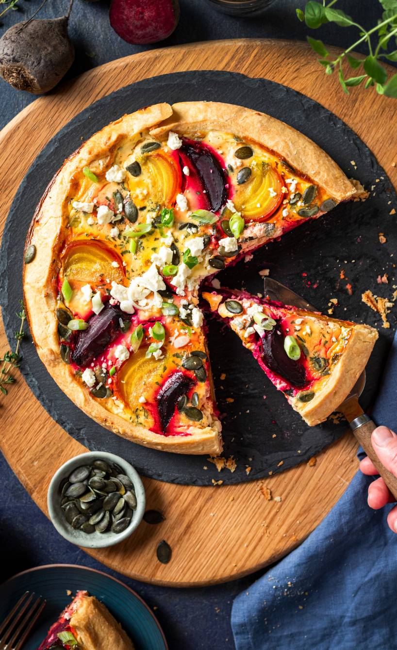 Runde Quiche mit roter und gelber Bete, Feta und Kürbiskernen angeschnitten auf einer Schieferplatte von oben