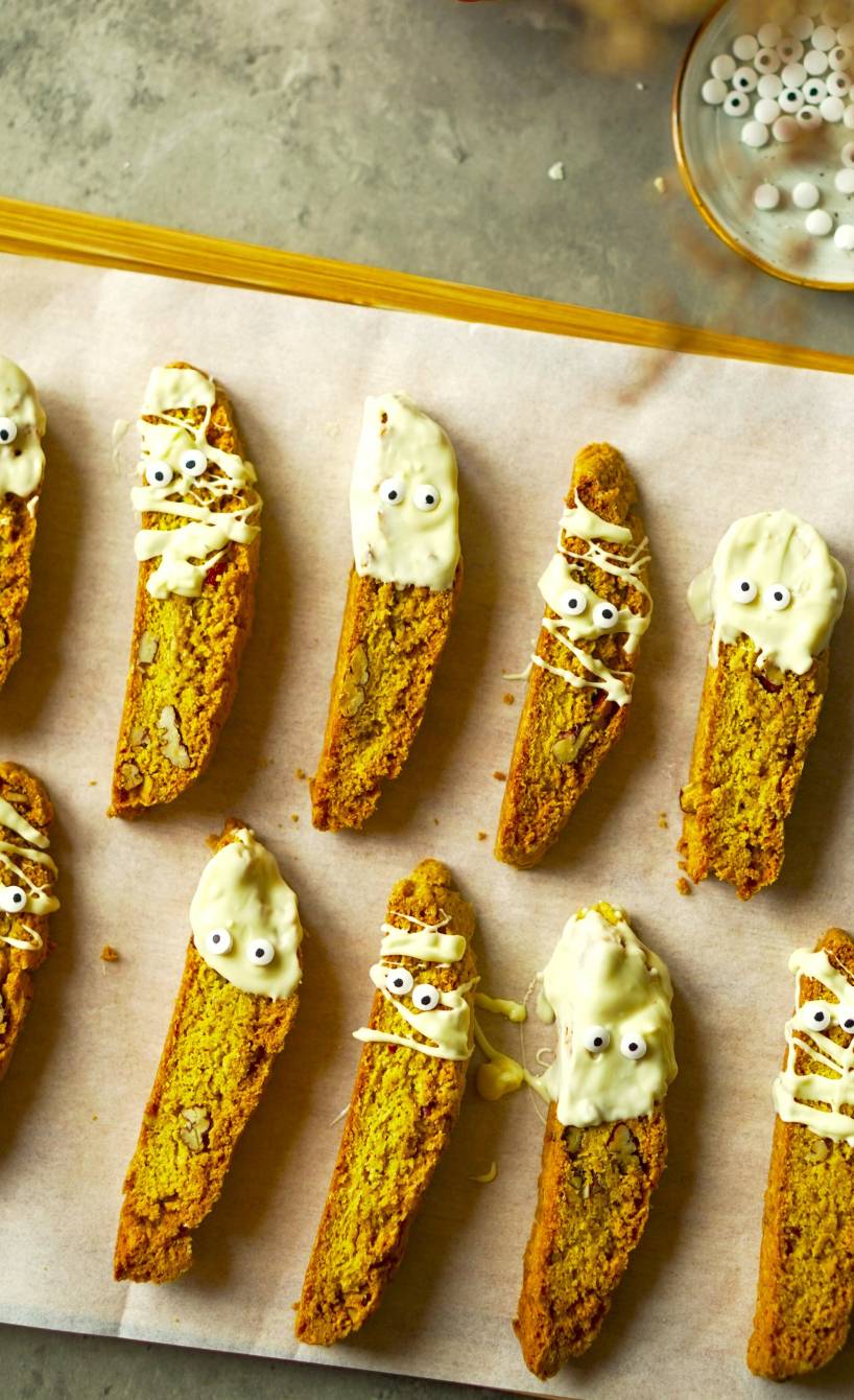 Mehrere Kürbis-Cantuccini nebeneinander auf einem Backblech dekoriert mit weißer Schokolade und Zuckeruagen als Mumien-Kekse