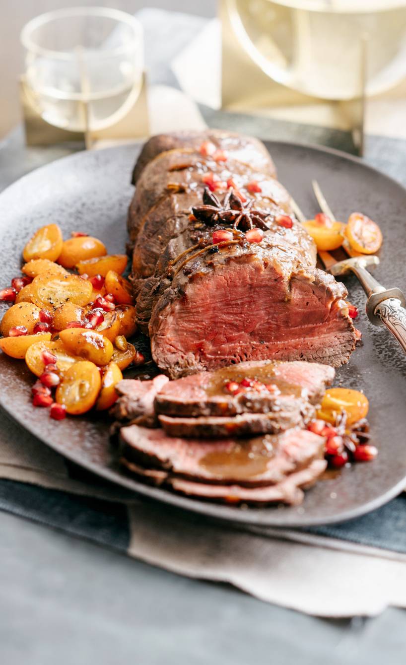 Rinderbraten angeschnitten auf einer Platte garniert mit karamelliserten Früchten, daneben Rotweinsoße