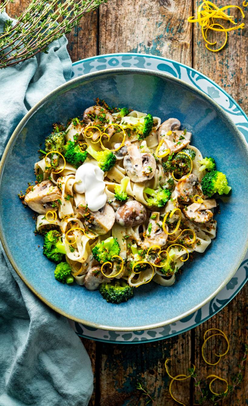 Tagliatelle in Champignon-Sahne-Sauce mit Brokkoli angerichtet auf einem tiefen Teller getoppt von Würzbröseln und Zitronenschalenstreifen